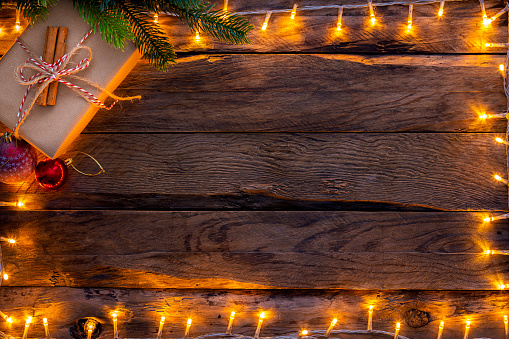Overhead view of Christmas string lights arranged all around the border of a rustic wooden table. A gift box is at the top left under a pine treee branch and complete the composition. Copy space ath the center of the frame.