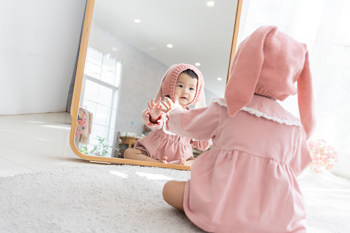 Rear view of little Asian baby girl wearing pink dress and headwear, sitting on soft carpet in front of mirror to see and play with her reflection image in mirror. Asian toddler girl wondering and feel curious about her image in the mirror in living room at home.