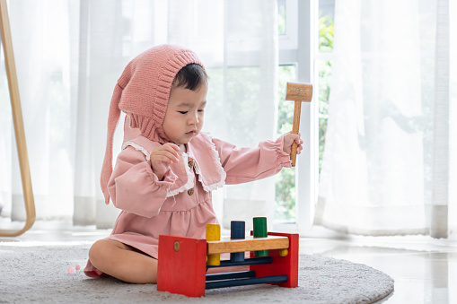 Cute Asian infant baby girl wearing pink dress and headwear sitting on soft carpet in living room, playing with multi-color wooden hammer block toy at home. Little girl having fun with colorful wooden toy.  Child development and learning.