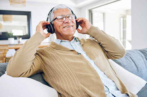 Senior man, headphones and listening to music in home, living room and relax on lounge sofa with podcast, radio or sound. Streaming, jazz song and audio with earphones and mobile technology in house