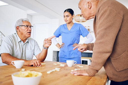 Nurse, senior and fighting over a game of dominoes with people in a retirement home for assisted living. Healthcare, medical and a female medicine professional looking at upset old men arguing