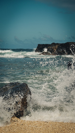 A vibrant blue wave crashing against jagged rocks along a sandy beach shoreline