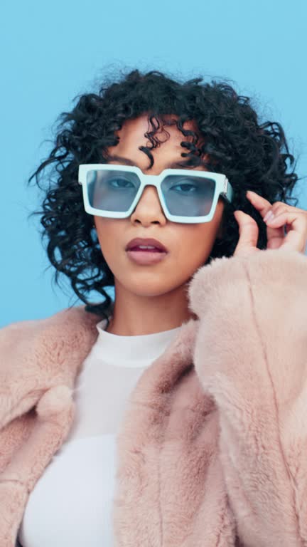 Fashion, woman and face with sunglasses, fur coat and model with confidence isolated on blue background. Style, trendy winter jacket and retro eyewear with accessory, portrait and serious in studio