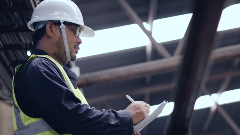 Mechanical maintenance engineer in protection workwear checking machine by clipboard in heavy factory, repairing check list