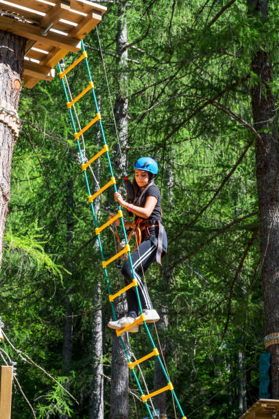 Happy young woman climbing ropes ladder in forest adventure park Side view of excited young female in helmet looking down while climbing hanging ladder from wooden bench with safety cables in sunlight during adventure trip in forest park girl climbing ladder stock pictures, royalty-free photos & images
