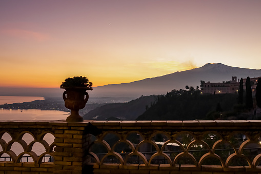 Scenic view on snow capped Mount Etna volcano and Mediterranean sea during sunset from public garden Parco Duca di Cesaro to Giardini Naxos in Taormina, Sicily, Italy, Europe, EU. Silhouette railing