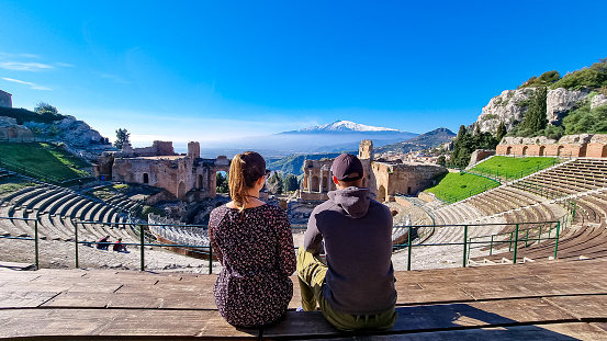 Tourist couple sitting with panoramic view on snow capped Mount Etna volcano on a sunny day seen from ancient Greek theater of Taormina, island Sicily, Italy, Europe, EU. Beautiful travel destination