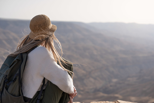 Rear view of tourist blonde woman with backpack and hat in viewpoint landscape in golden hour of wilderness Canyon Wadi Mujib in Jordan
