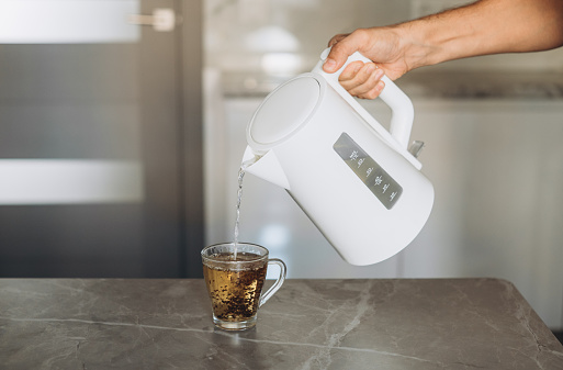 Male hands pouring water from a white electric kettle into a mug for making tea