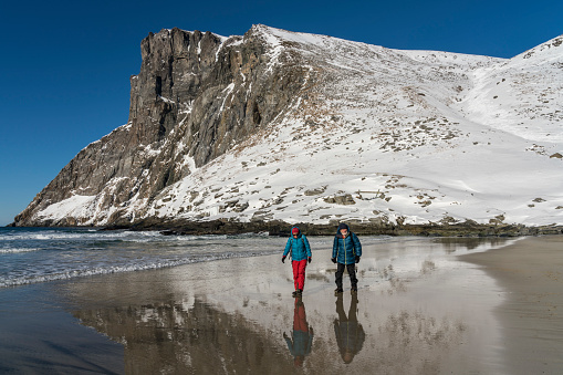Man and women walking on the Kvalvika Beach, they are reflecting in the sea in winter. Close to the Moskensoynear fishing village Fredvang on the Lofoten Islands. Norway.