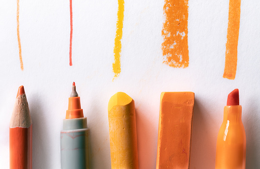 An Orange Pencil, Pen, Pastels, and Marker Drawing lines on White Background