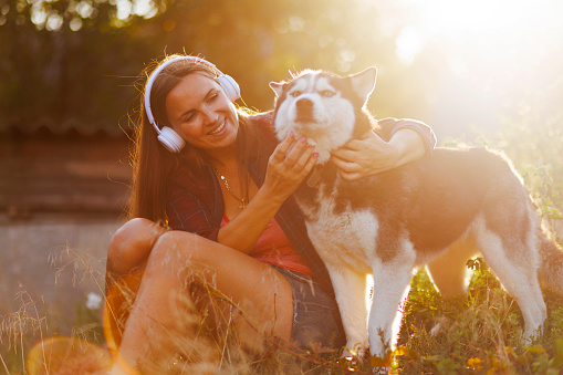 Beautiful young woman in headphones plays, hugs with her dog outdoors at sunset.
