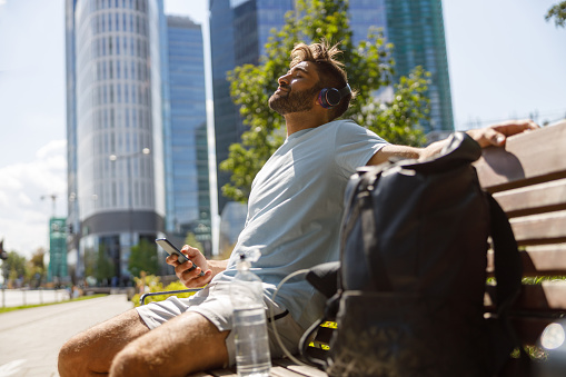 Male tourist resting sitting on bench and listen music in headphones on skyscrapers background