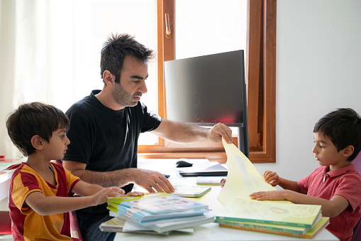 Father binding his son's books at his desk. Father and son are preparing for school together. There is a whiteboard in the foreground. It was shot with a full frame camera in daylight in the study room.