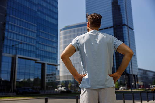 Back view of handsome sporty man standing in modern big city and looking on glass skyscrapers