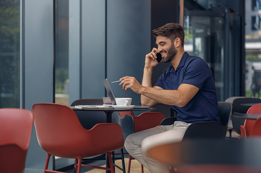 Handsome male freelancer talking on phone with client while working on a laptop in coworking