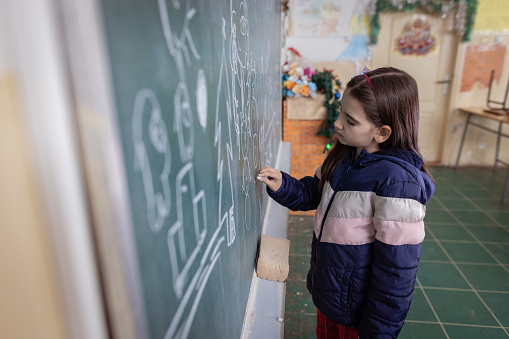 A rear view of a little cute girl standing in the classroom in front of a chalkboard and drawing Christmas ornaments with chalk.