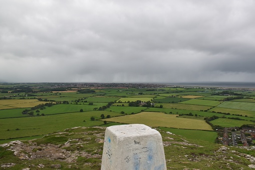 View across patchwork farm fields from above with a trig point in the foreground against cloudy overcast sky