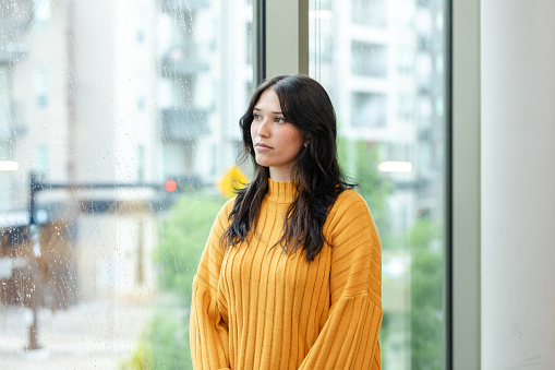 A beautiful young woman looks through a window with a worried look on her while standing in her city apartment.