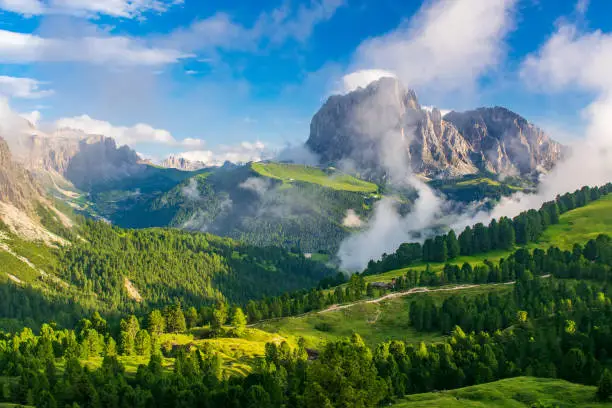 A vibrant depiction of the Sassolungo Massif and Gardena Valley in Dolomite Alps, Italy. The foreground showcases a lush green valley, while the background highlights a cloud-covered mountain peaks