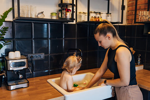 In a modern apartment, a caring mother bathes her baby girl in the kitchen sink, while she has fun with her toys