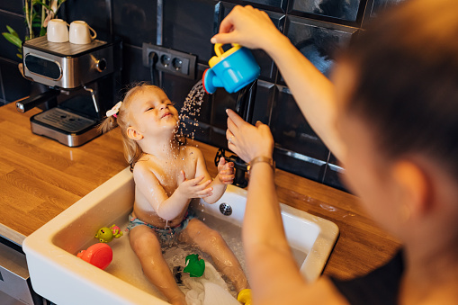 A caring mother bathes her cute little baby girl in the kitchen sink, while she has fun with her toys