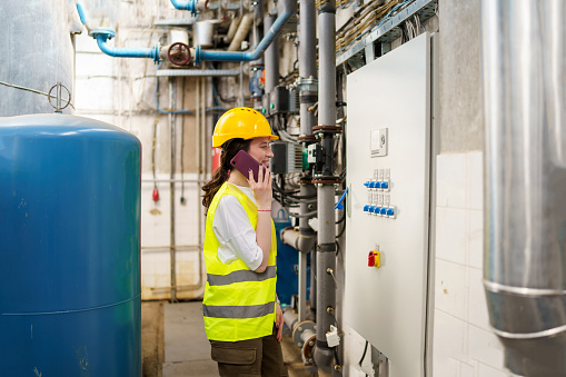 A young woman in a hard hat and safety vest multitasks while using her mobile phone to communicate with her colleagues and document her work in a heating plant