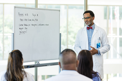 A mature adult male doctor uses a whiteboard when he teaches an education training class to a unrecognizable group of medical professionals.
