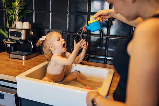 A mother splashes her cheerful baby girl with a watering can while the baby is bathed in the kitchen sink