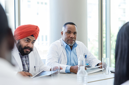 During the meeting in the hospital board room, two diverse male doctors listen attentively to an unseen colleague.