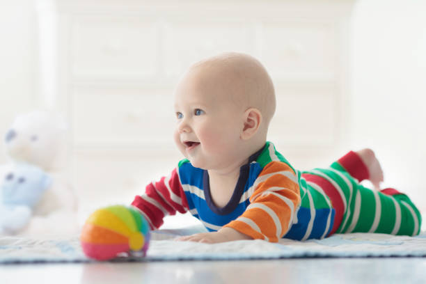 Baby boy with toys and ball Adorable baby boy learning to crawl and playing with colorful rainbow ball toy in white sunny bedroom. Cute laughing child crawling on a play mat. Nursery, clothing and toys for little kids. bear stomach stock pictures, royalty-free photos & images