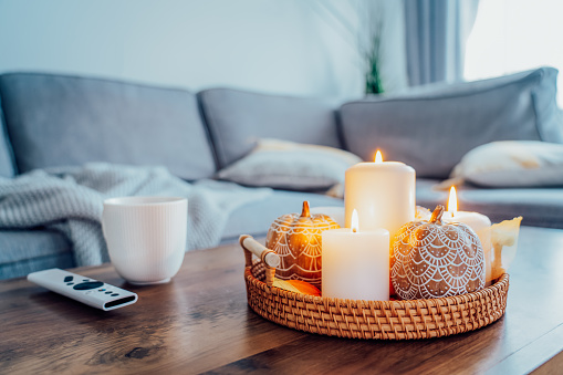 Autumn fall cozy mood composition for hygge home decor. Orange pumpkins, burning candles, cup with hot drink and remote controller on coffee table in living room. Movie night at home. Cozy relax time