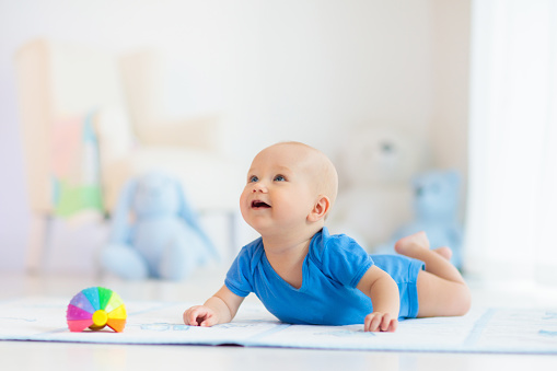 Adorable baby boy learning to crawl and playing with colorful rainbow ball toy in white sunny bedroom. Cute laughing child crawling on a play mat. Nursery, clothing and toys for little kids.