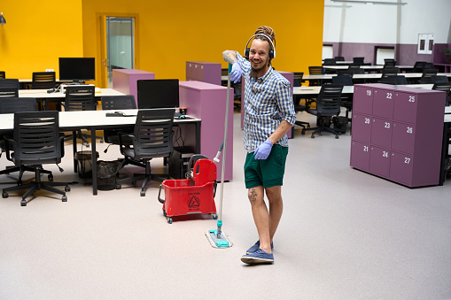 Cheerful cleaner is informal in a coworking space with work gadgets, he cleans the office space