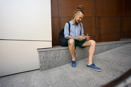 Smiling freelancer chatting online on the phone on a granite curb near a modern building