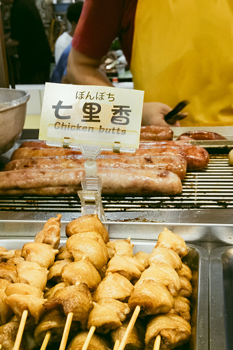 A grilled meat vendor with a funny sign sells chicken butts and sausages at a crowded and busy night market in Taipei, Taiwan.  Diners are seated in the background.