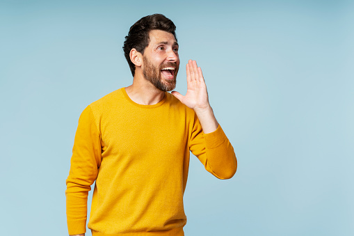 Excited handsome bearded man holding hand near face, screaming loud looking away isolated on blue background. Emotional hipster doing sales announcement. Promotion, shopping concept