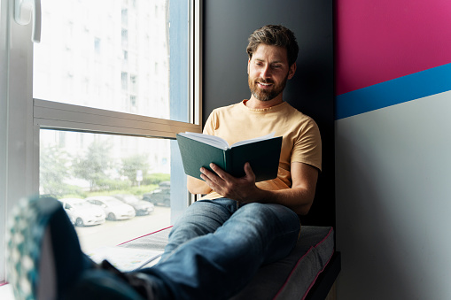 Handsome smiling bearded man reading book sitting at home near window. Smart middle aged student studying, learning language, exam preparation in university campus. Education concept