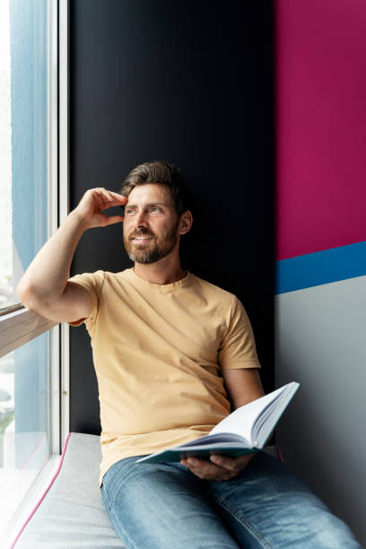 istock Smiling man reading book sitting at home looking at window. Student studying, learning language 1681429773