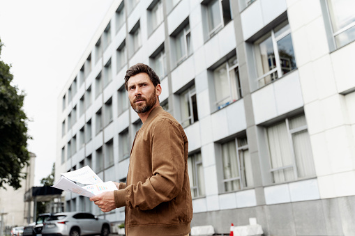 Portrait of handsome serious businessman, worker wearing stylish leather jacket holding paper documents looking away standing near office building, copy space. Successful business concept