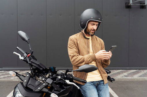 Stylish confident man, biker wearing helmet using mobile phone standing near sport motorcycle. Handsome fashion model wearing stylish leather jacket posing for pictures on motorbike