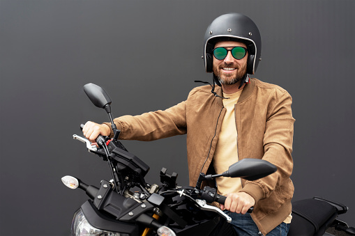 Portrait of handsome smiling bearded man, biker wearing helmet, stylish eyeglasses, leather jacket riding motorcycle isolated on black background. Journey, freedom, transportation concept