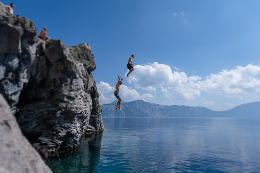 Profile view of a fit young man and his adventurous senior adult father-in-law jumping off a cliff into Crater Lake on a warm and sunny summer day in Oregon.