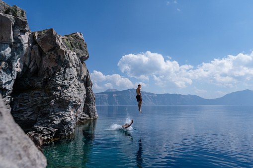 Profile view of a fit young man and his adventurous senior adult father-in-law jumping off a cliff into Crater Lake on a warm and sunny summer day in Oregon.