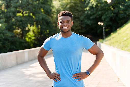 Portrait of attractive smiling African American man in blue t shirt posing for picture, looking at camera. Handsome athlete training outdoors in park. Concept of motivation, sport, healthy lifestyle