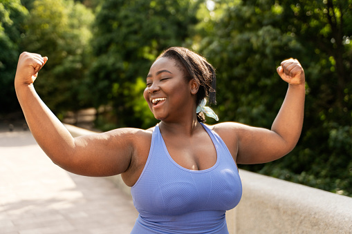 Portrait of beautiful smiling African American woman in sportswear showing biceps exercising on the street, outdoors, looking away. Attractive female posing for picture. Body transformation concept
