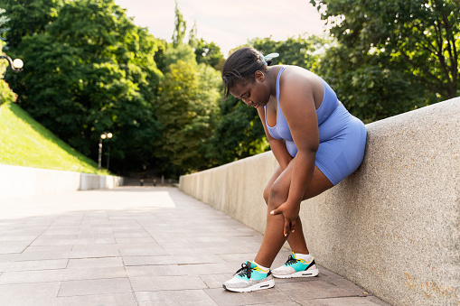 Pensive attractive african american man in sportswear and sneakers exercising in park, holding knee, pain, trauma, copy space. Plus size young female. Health care concept