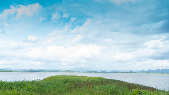 Ground covered with green grass juts out into the water. Large reservoir surrounded by mountains Under blue sky and white clouds. Pra Sae Reservoir Rayong Thailand.