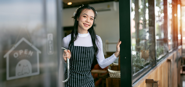 Cheerful young waitress waiting for clients at coffee shop. Successful small business owner in blue apron standing at entrance