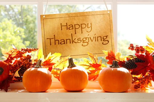 A Happy Thanksgiving sign hangs in a window as several pumpkins and a fall garland of autumn leaves rest on the window sill as a late afternoon sun filters through the window.